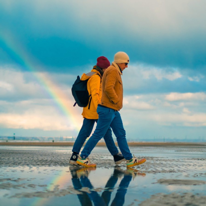 Relaxed couple walking on a low tide beach on a cold day, holding hands, a rainbow in the background