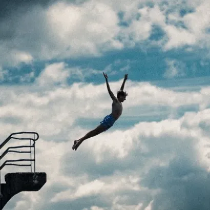 Teenage boy jumping from a high diving board, stretching out to a lightly cloudy sky