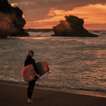 Middle aged person carrying a surf board under his arm, reflecting, looking at the the sea and waves, an orange sunset in the background.